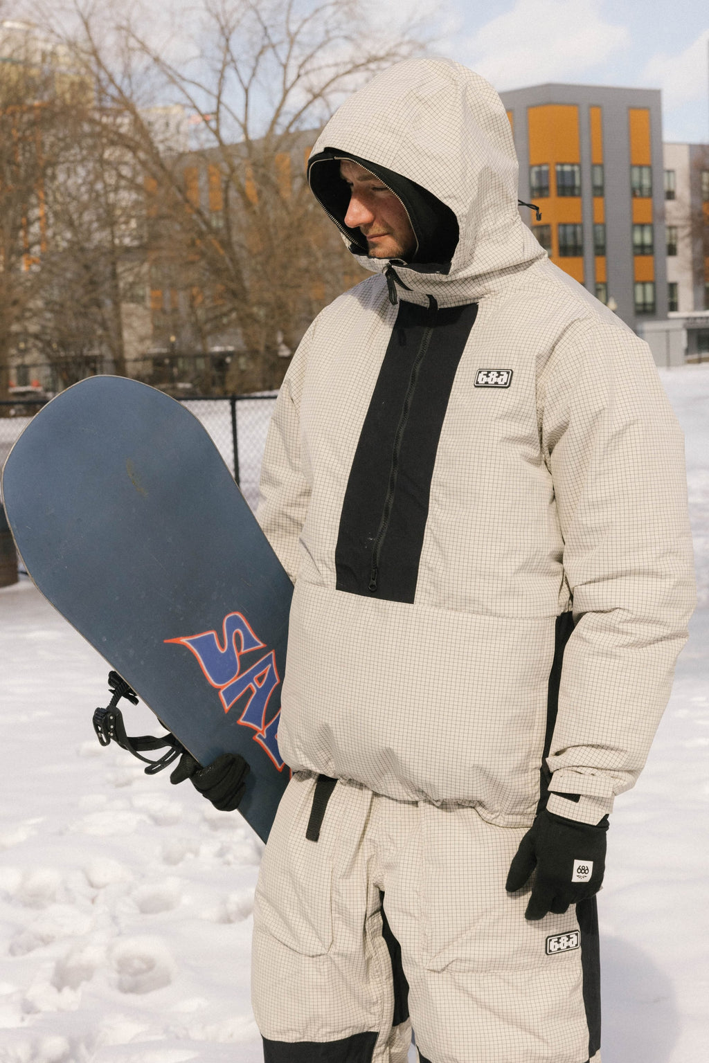 A person stands in the snow holding a snowboard, wearing the 686 Men's Ghost™ 2.5L Anorak. Trees and modern buildings are visible in the background.