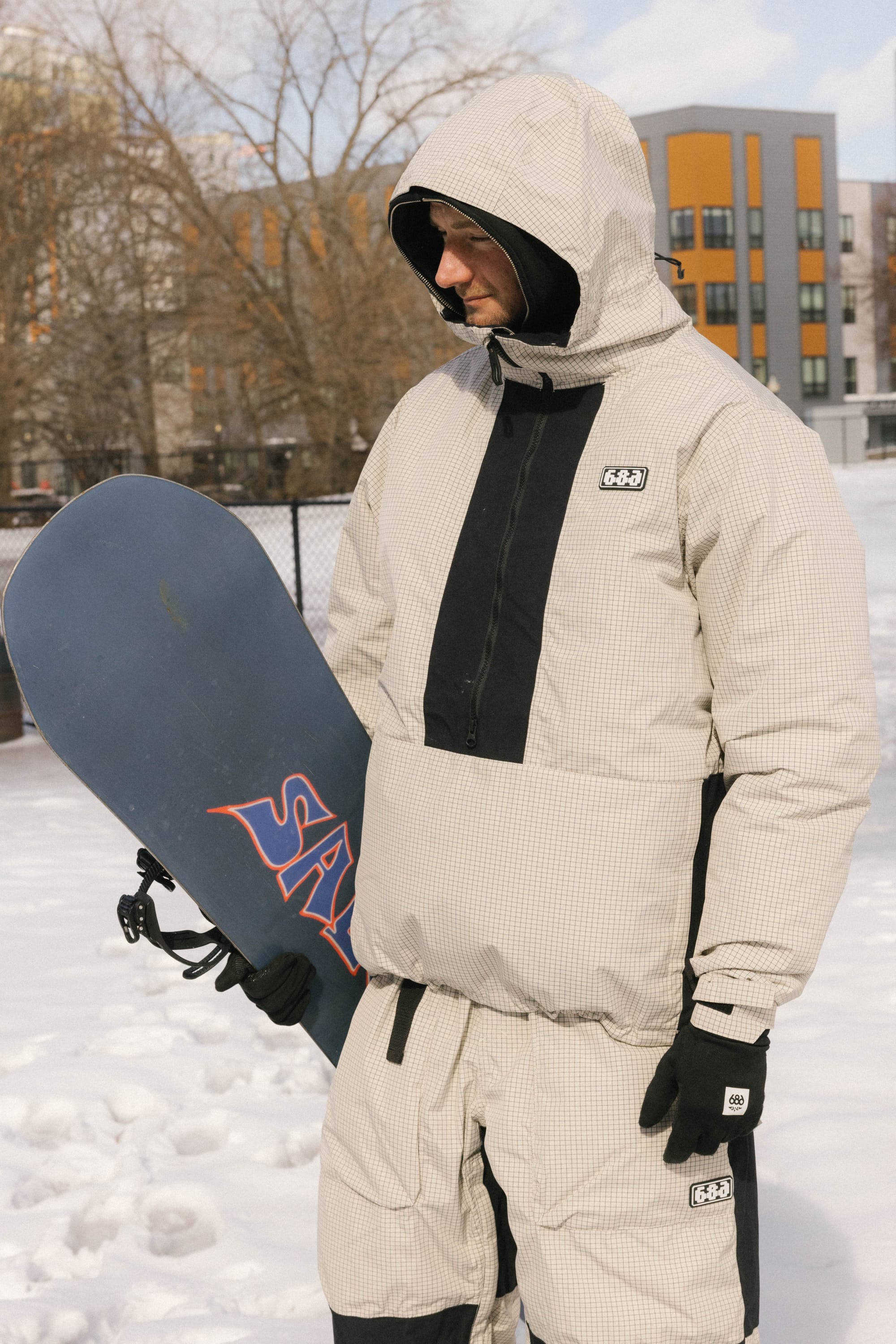 A person stands in the snow holding a snowboard, wearing the 686 Men's Ghost™ 2.5L Anorak. Trees and modern buildings are visible in the background.