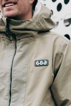 A person wears the 686 Men's Ghost™ 2.5L Anorak—a tan, rugged nylon hooded jacket with a pixel-style chest badge—by 686, standing before a white wall with circular cutouts. Only the lower half of their face is visible.