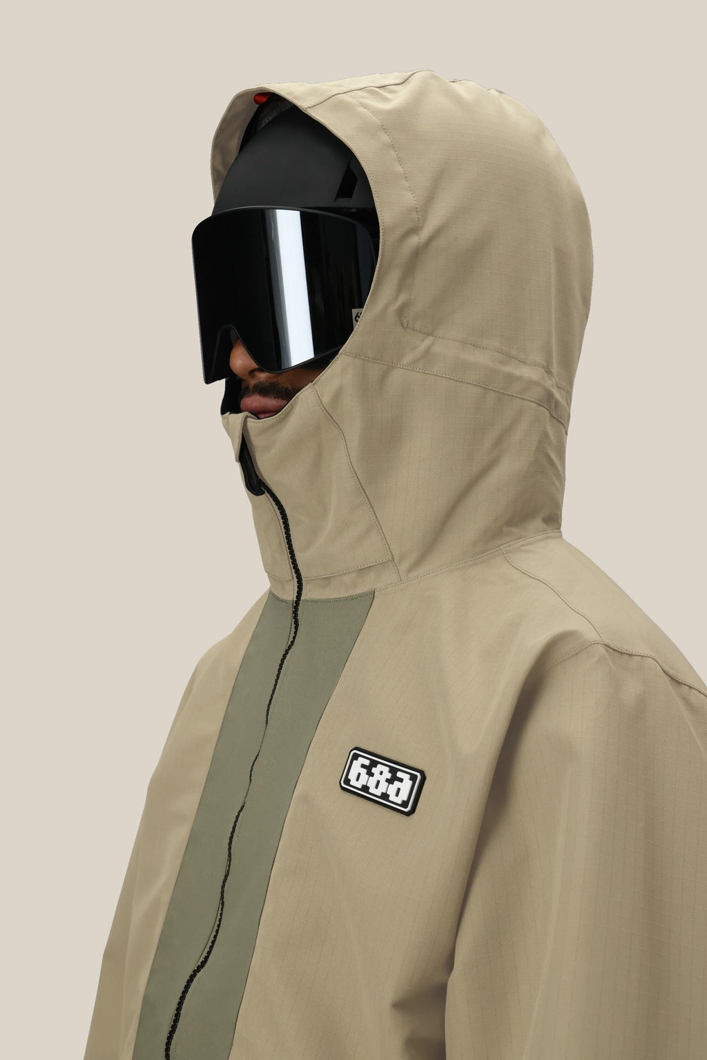 A man wearing the 686 Men's Ghost™ 2.5L Anorak in beige, featuring a 686 patch, and large black ski goggles, is shown in profile against a plain light background.