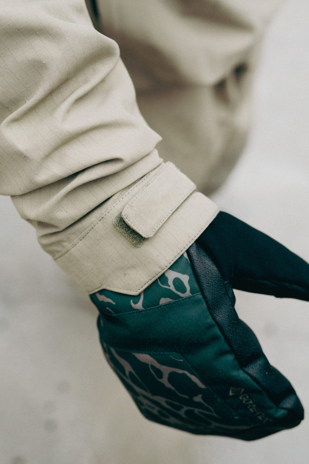 A close-up of a person's arm wearing the 686 Men's Ghost™ 2.5L Anorak in beige, featuring a rugged nylon build with a Velcro wrist strap and paired with a black and green patterned glove.