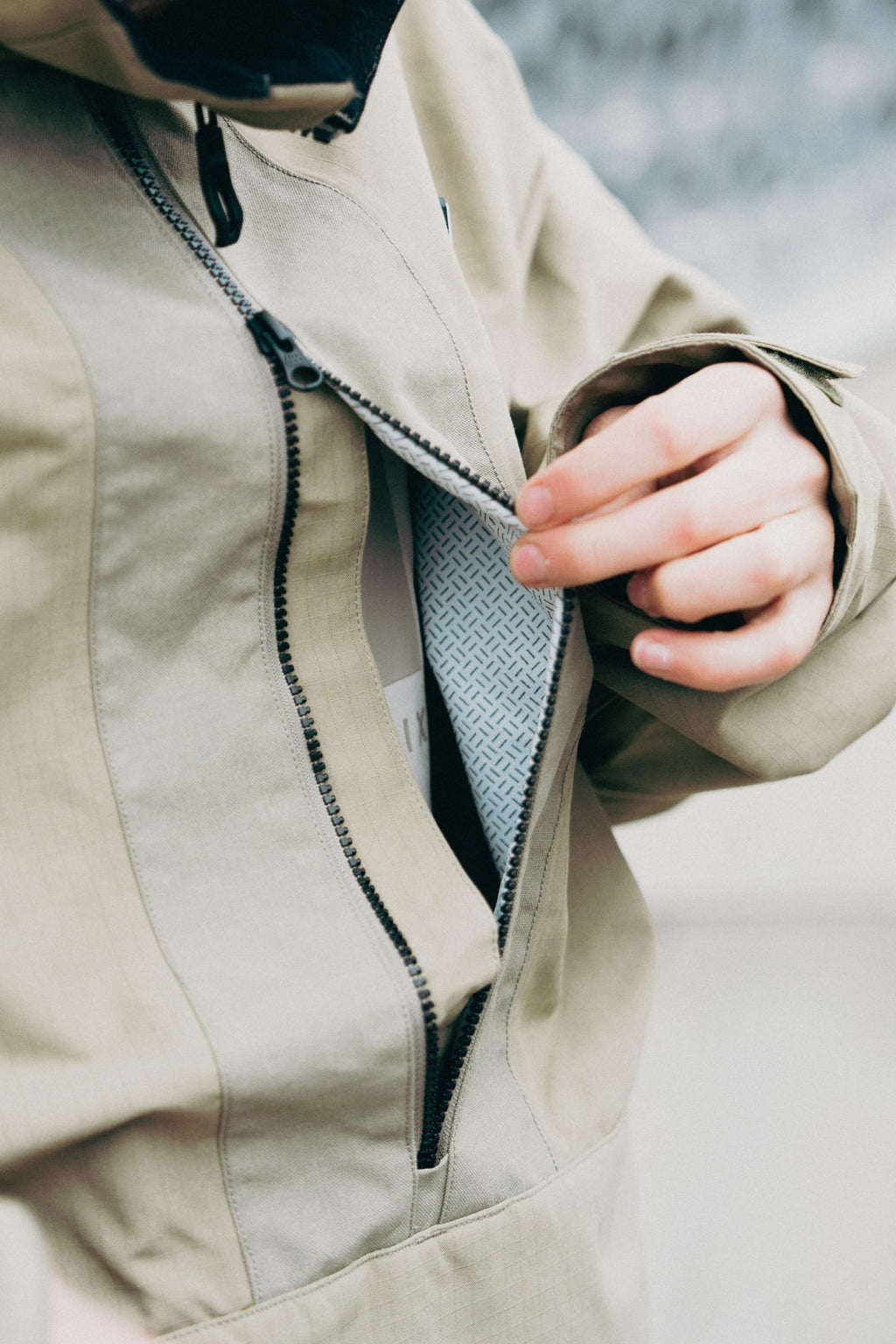 A hand unzips the 686 Men's Ghost™ 2.5L Anorak in beige, showcasing its rugged nylon construction and revealing a patterned lining over a white layer, with a softly blurred background.