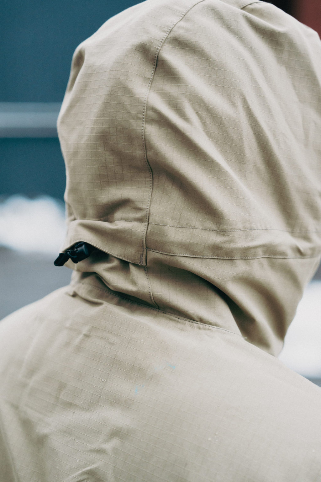 A close-up of a person wearing the 686 Men's Ghost™ 2.5L Anorak in beige, seen from behind with the hood up and secured by a black drawstring toggle. The blurred background features white areas that may be snow.