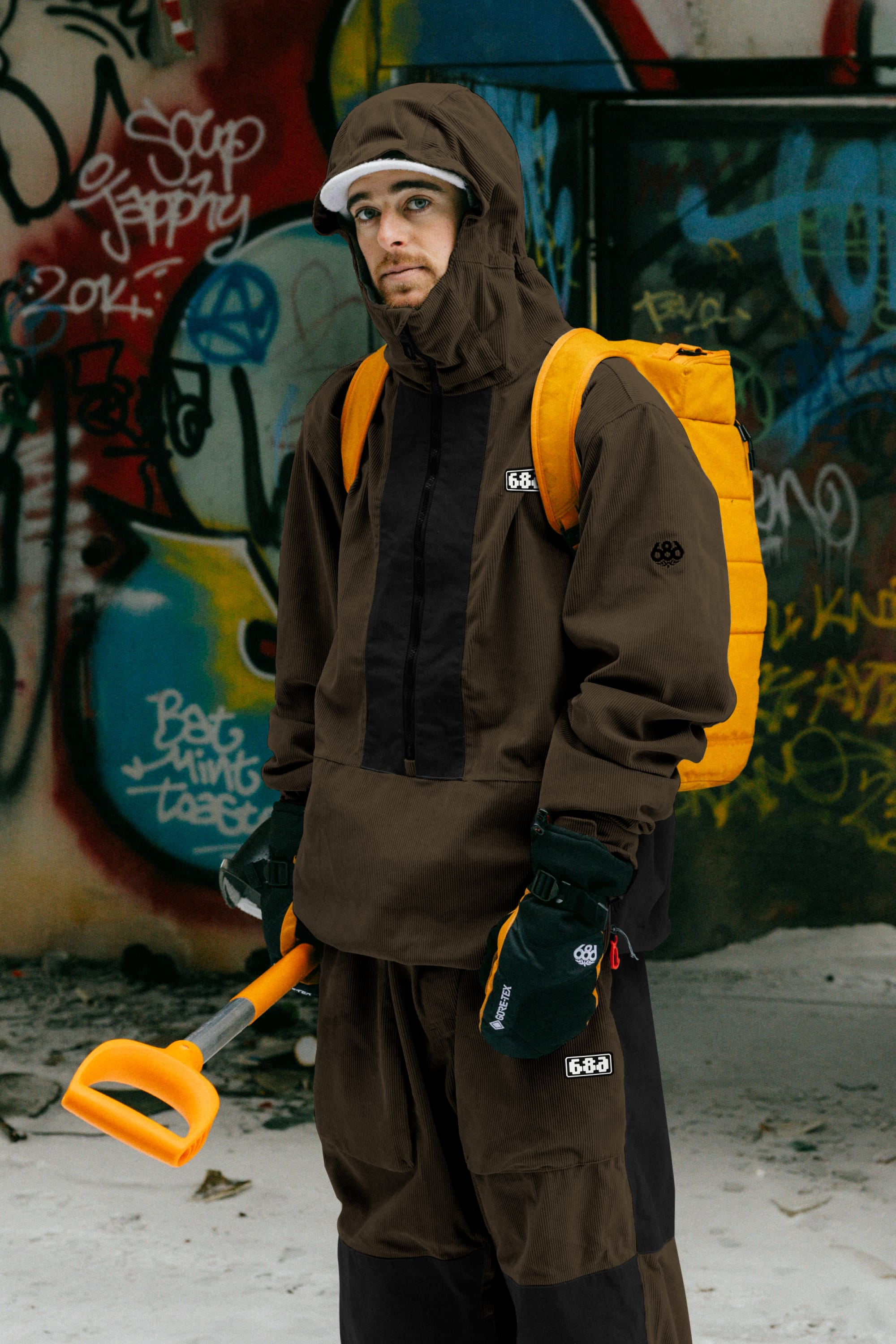 Wearing 686 Ghost™ 2.5L Pant by 686, a person in brown winter gear and gloves stands on snow with an orange backpack and shovel, against graffiti-covered walls. The pants offer flexibility and ruggedness for winter adventures.