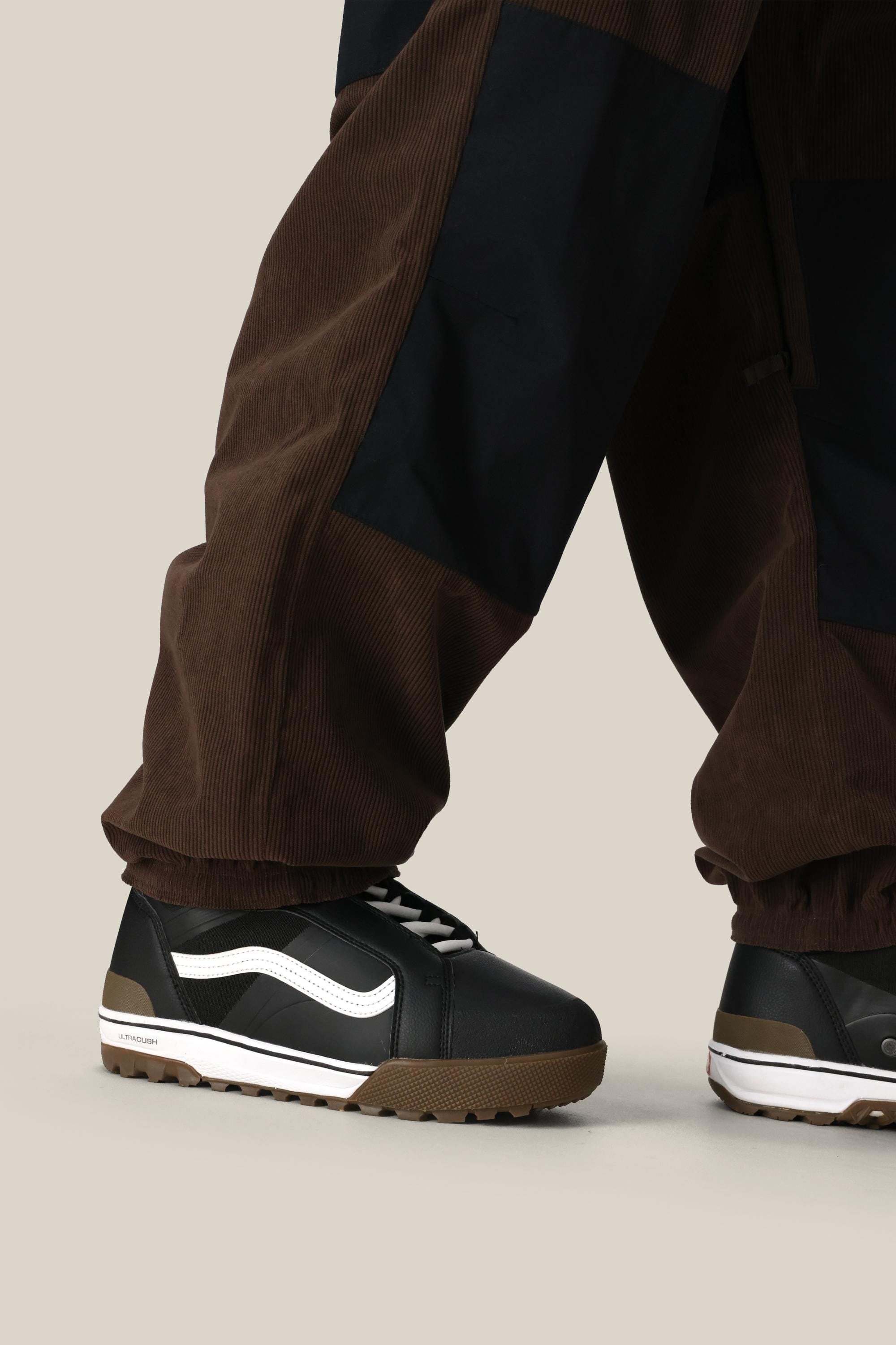 Someone wearing the 686 Ghost™ 2.5L Pant and black sneakers with white detailing and a brown sole stands on a neutral beige background.