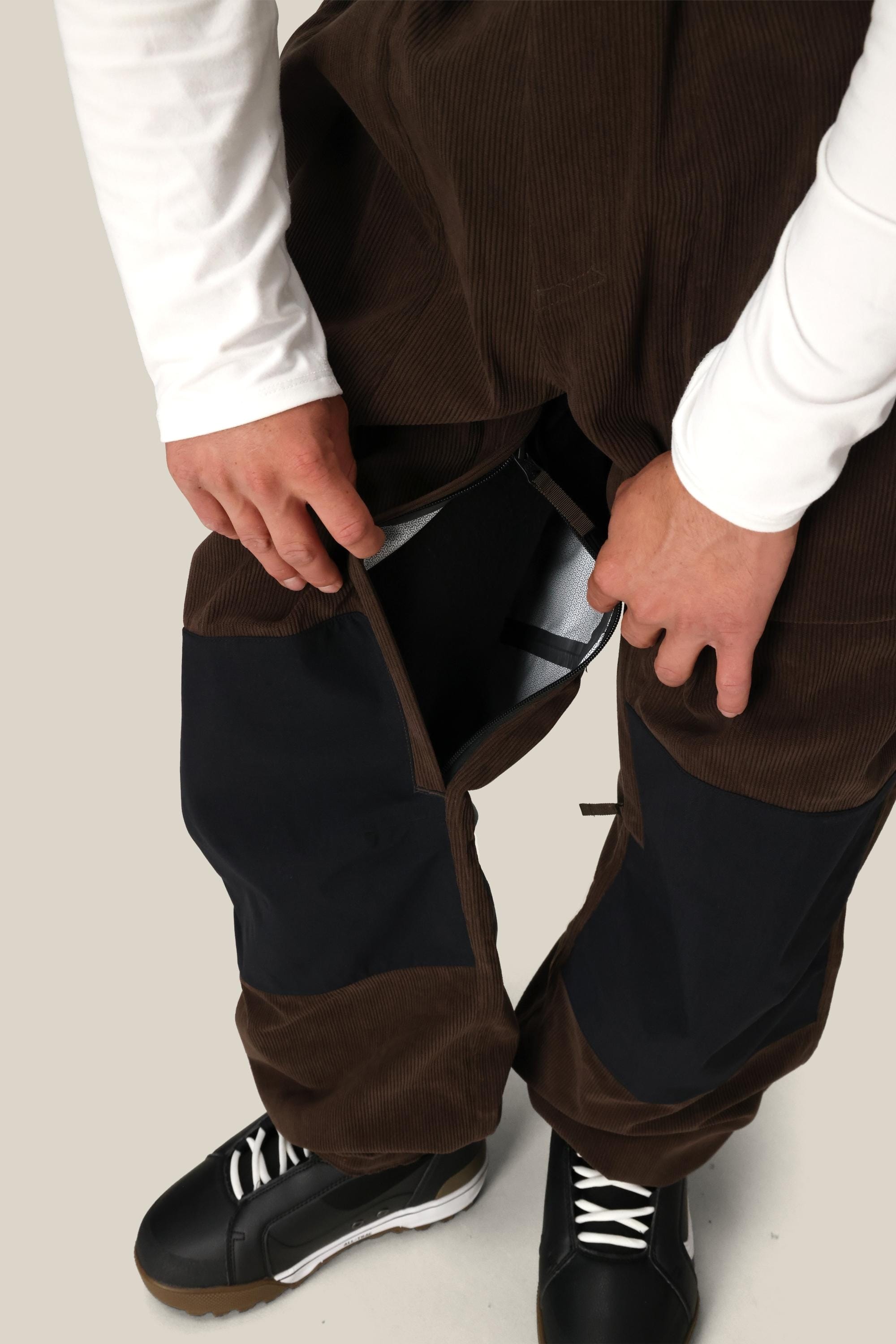 A person wearing 686 Ghost™ 2.5L Pants with zippered vents pulls one vent open to show the inside. They’re also dressed in a white long-sleeve shirt and black sneakers.