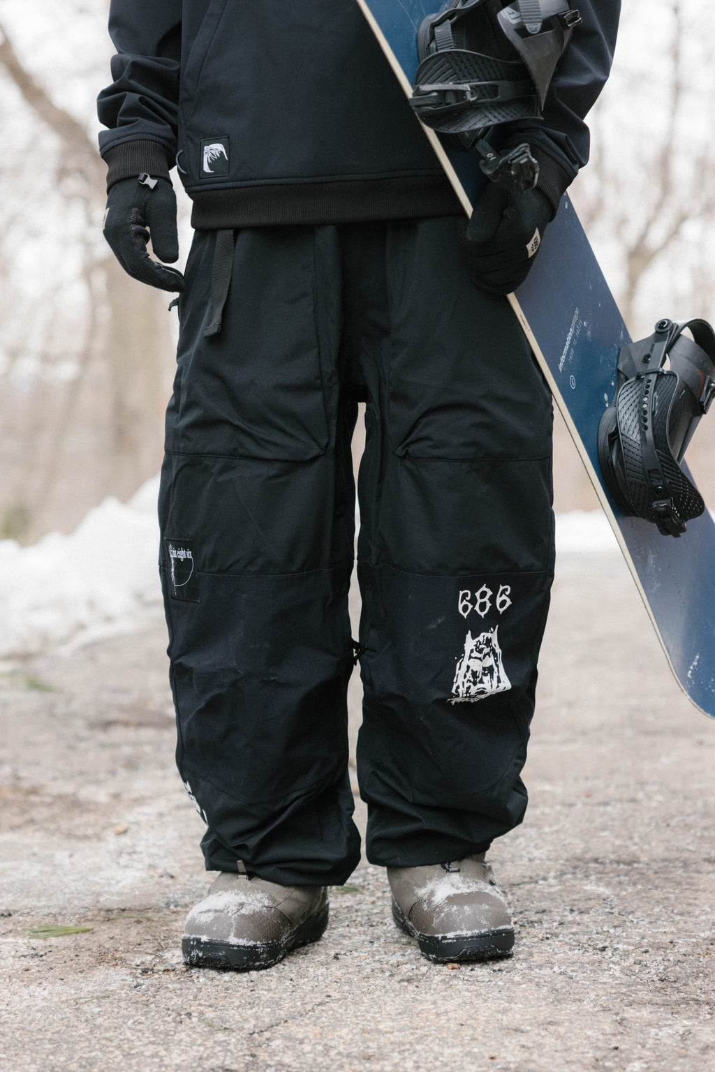 A person in 686 Ghost™ 2.5L Pants and boots stands on a snowy path, holding a snowboard with black bindings upright. Patches and graphics accent the pants against a wintry, wooded backdrop.