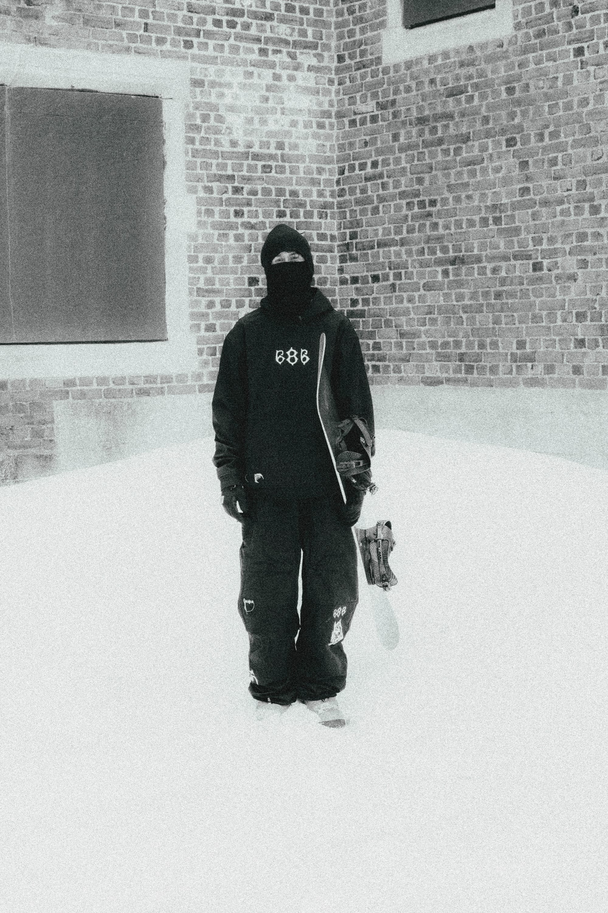 Wearing 686 Ghost™ 2.5L Pants, a person in dark winter gear and a ski mask stands on snow holding a snowboard, with a brick building behind them.