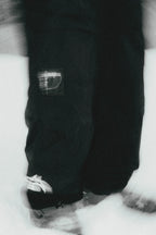 Black and white close-up of a person in 686 Ghost™ 2.5L Pants with snowboard boots, standing on snow. The “six eight six” patch is visible; the dynamic, slightly blurred effect highlights the rugged style from 686.