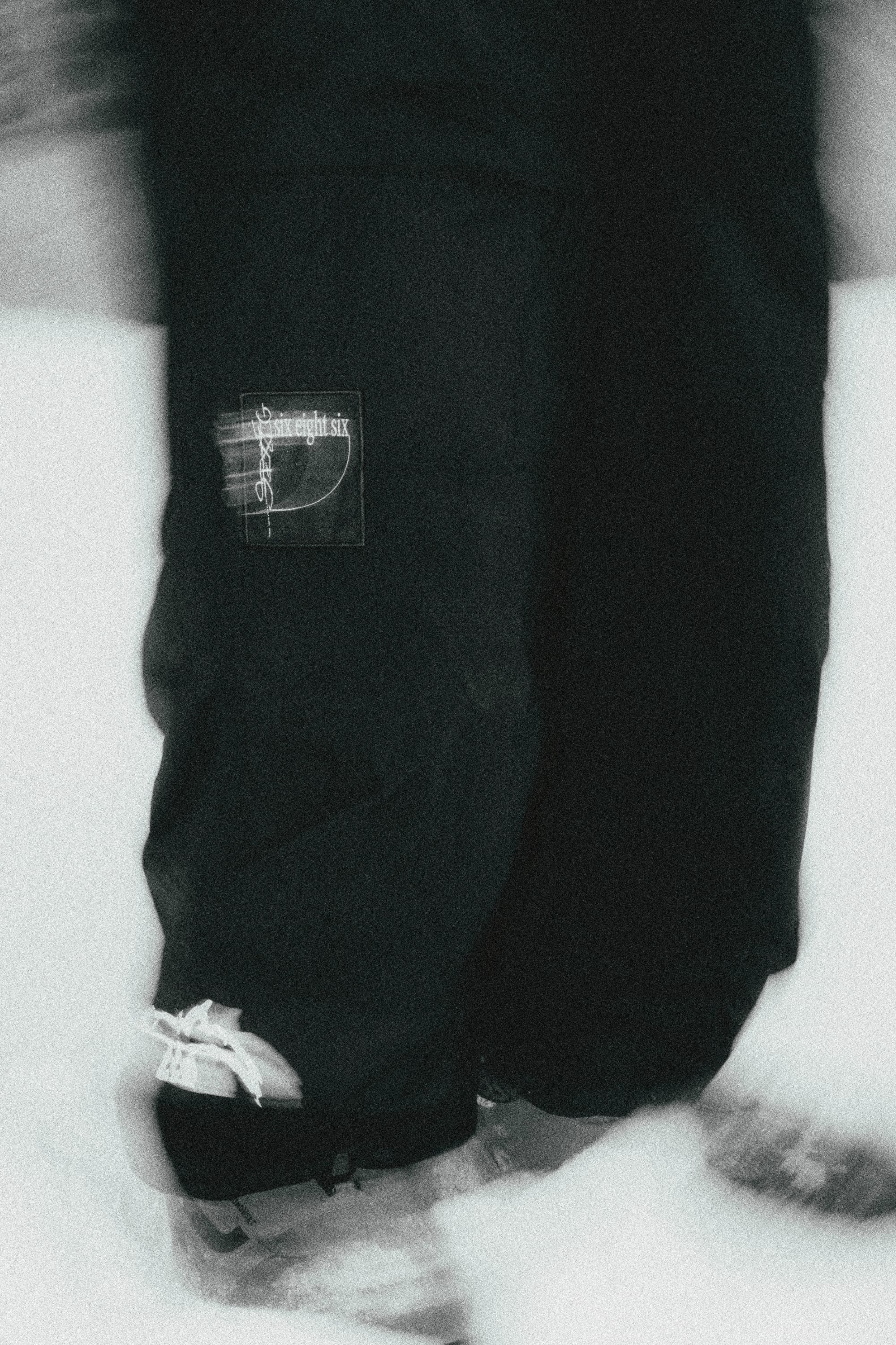 Black and white close-up of a person in 686 Ghost™ 2.5L Pants with snowboard boots, standing on snow. The “six eight six” patch is visible; the dynamic, slightly blurred effect highlights the rugged style from 686.