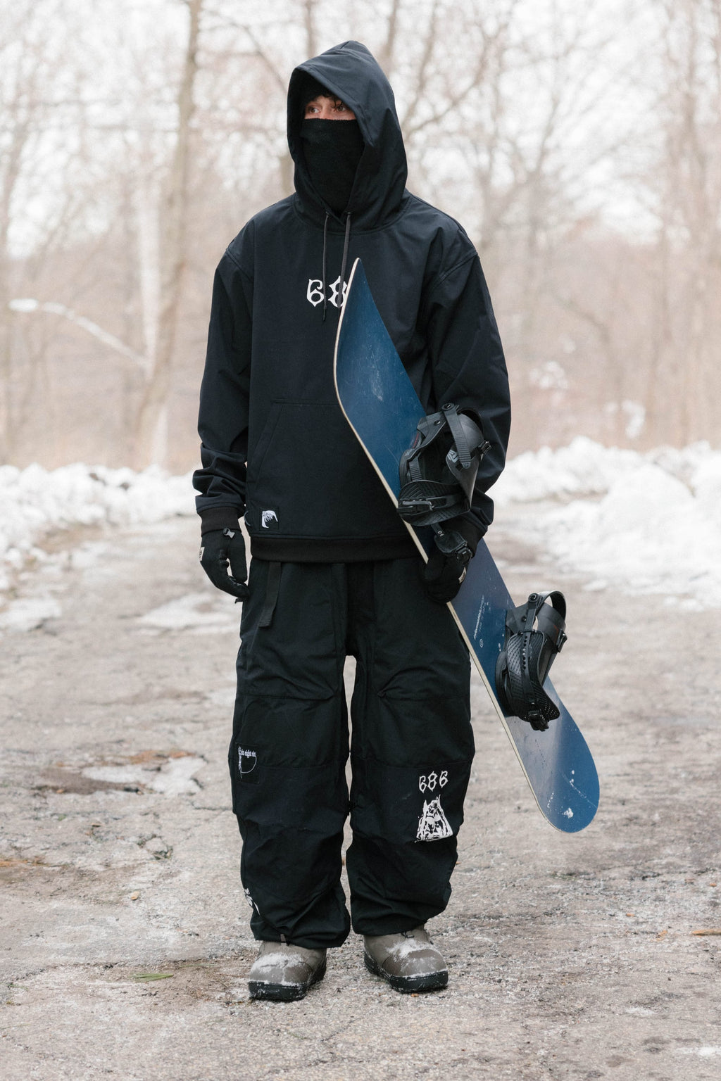 Dressed in black 686 Ghost™ 2.5L Pant and winter gear, a person stands on a snowy path holding a blue snowboard, with snow-covered trees in the background.