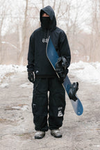 Dressed in black 686 Ghost™ 2.5L Pant and winter gear, a person stands on a snowy path holding a blue snowboard, with snow-covered trees in the background.