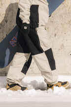 A person wearing 686 Ghost™ 2.5L Pants and winter boots walks on snow with gloves, holding a snowboard against a concrete wall.