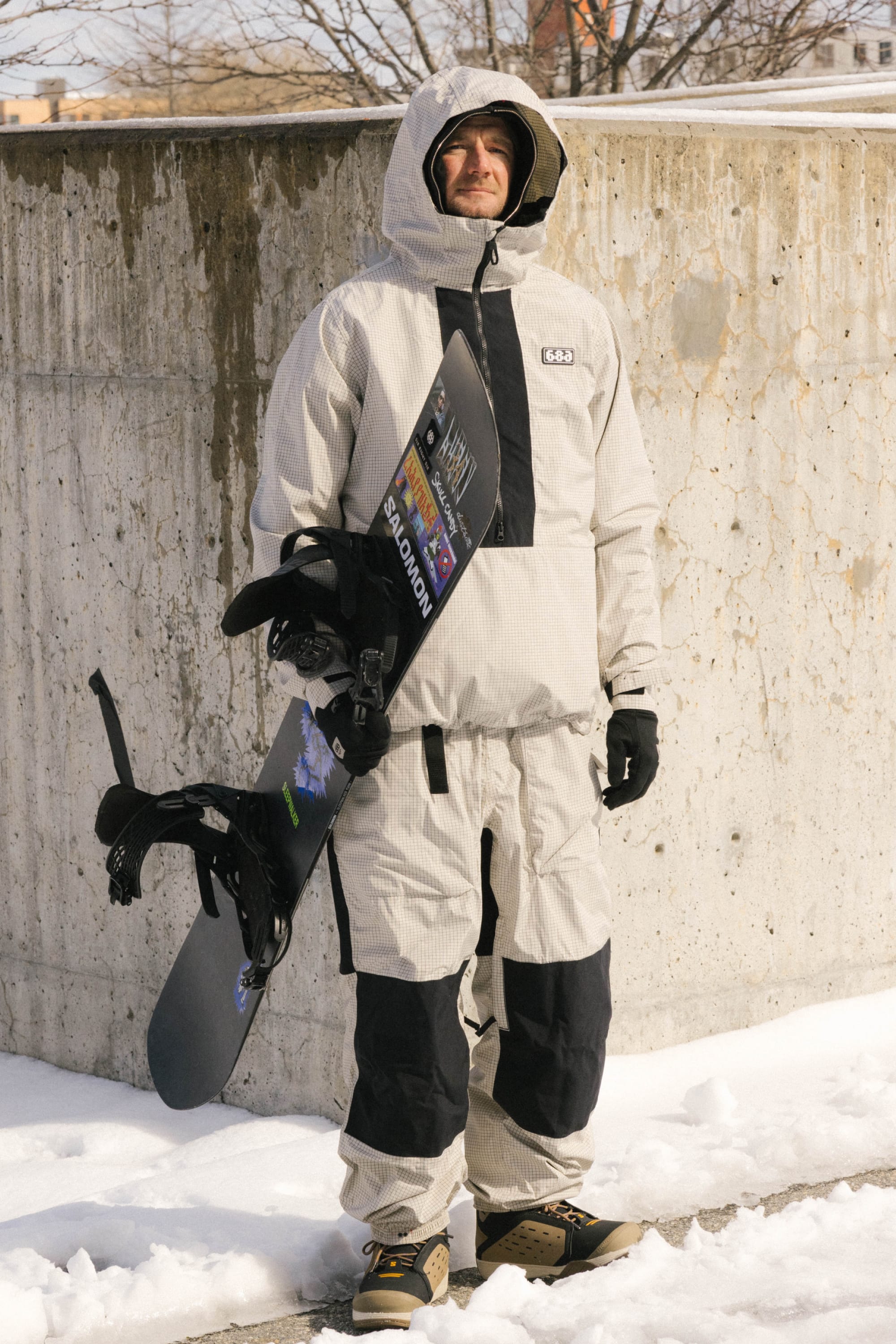 A person in a light gray and black snowsuit stands on snow holding a snowboard. They wear gloves, boots, and the flexible 686 Ghost™ 2.5L Pant. A concrete wall and bare trees are in the background.