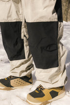 A close-up of a person standing on snow, wearing 686 Ghost™ 2.5L Pants with reinforced knees and tan, black, and yellow winter shoes.