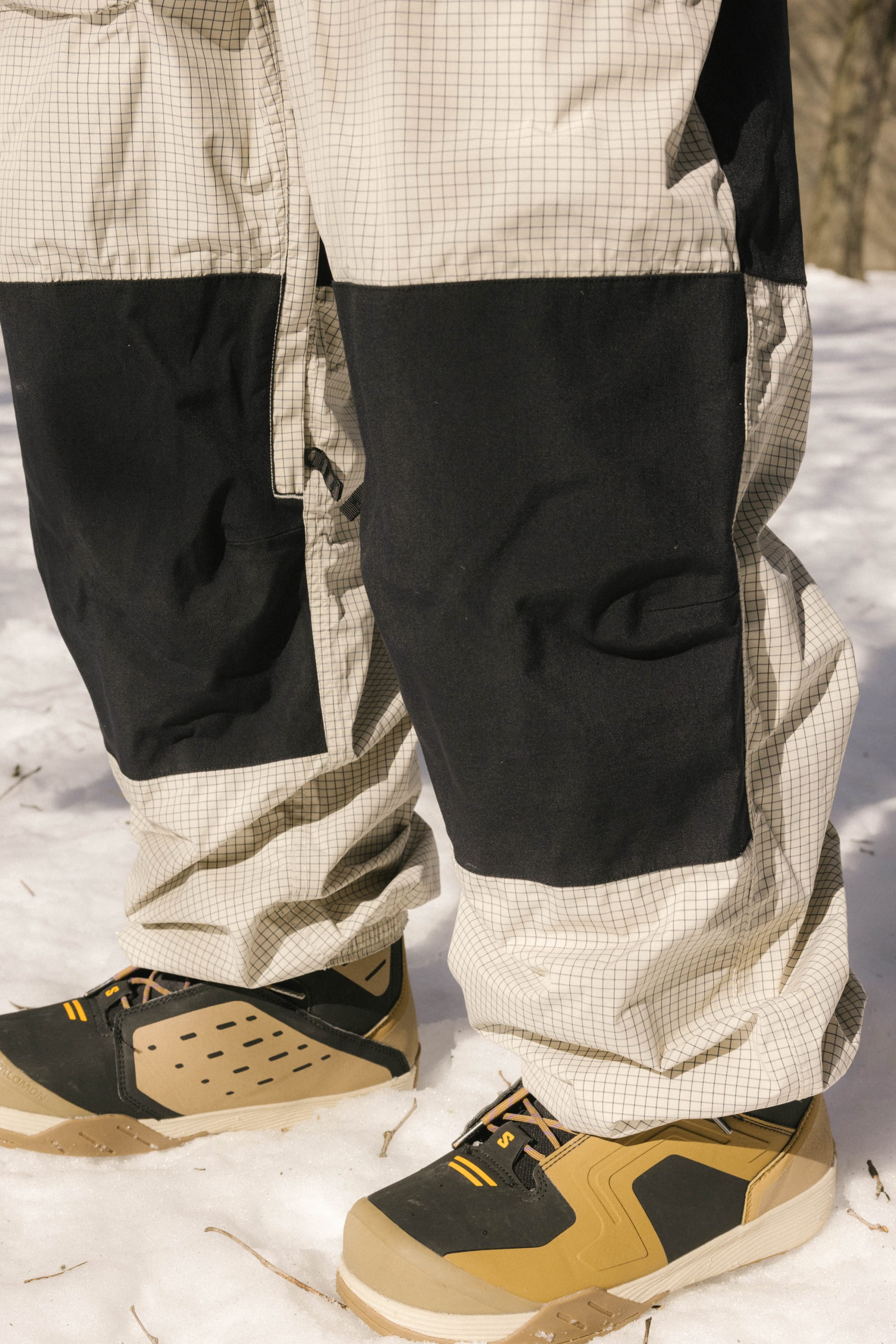A close-up of a person standing on snow, wearing 686 Ghost™ 2.5L Pants with reinforced knees and tan, black, and yellow winter shoes.