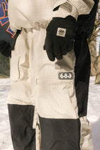 Wearing a beige and black checkered snowsuit with 686 patches, black gloves, and 686 Ghost™ 2.5L Pant, a person stands on snow outdoors holding a snowboard.