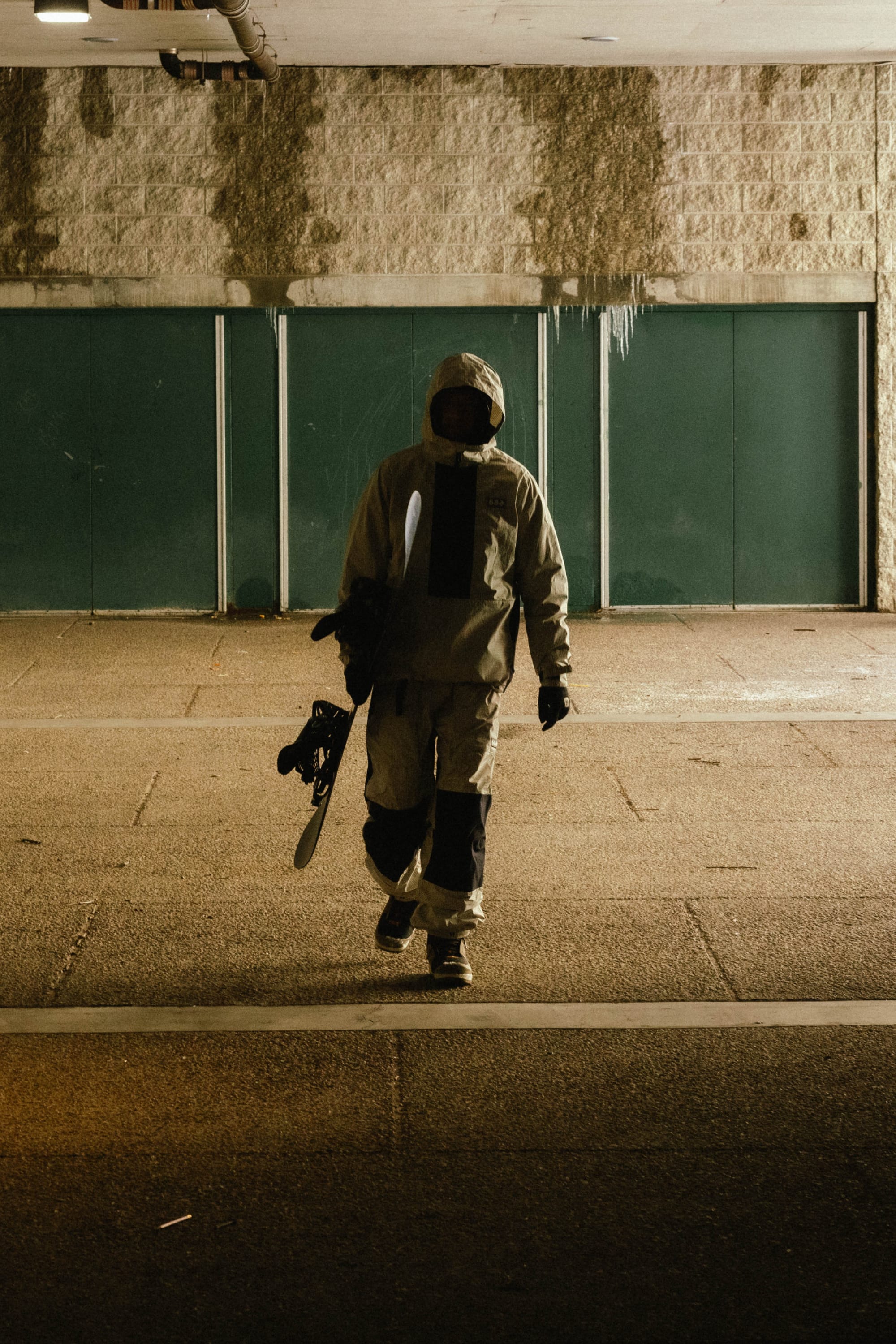 A person in a hooded jacket and mask walks indoors with a snowboard, against a concrete wall and green doors under dramatic lighting. They wear the 686 Ghost™ 2.5L Pant by 686 for comfort and style.