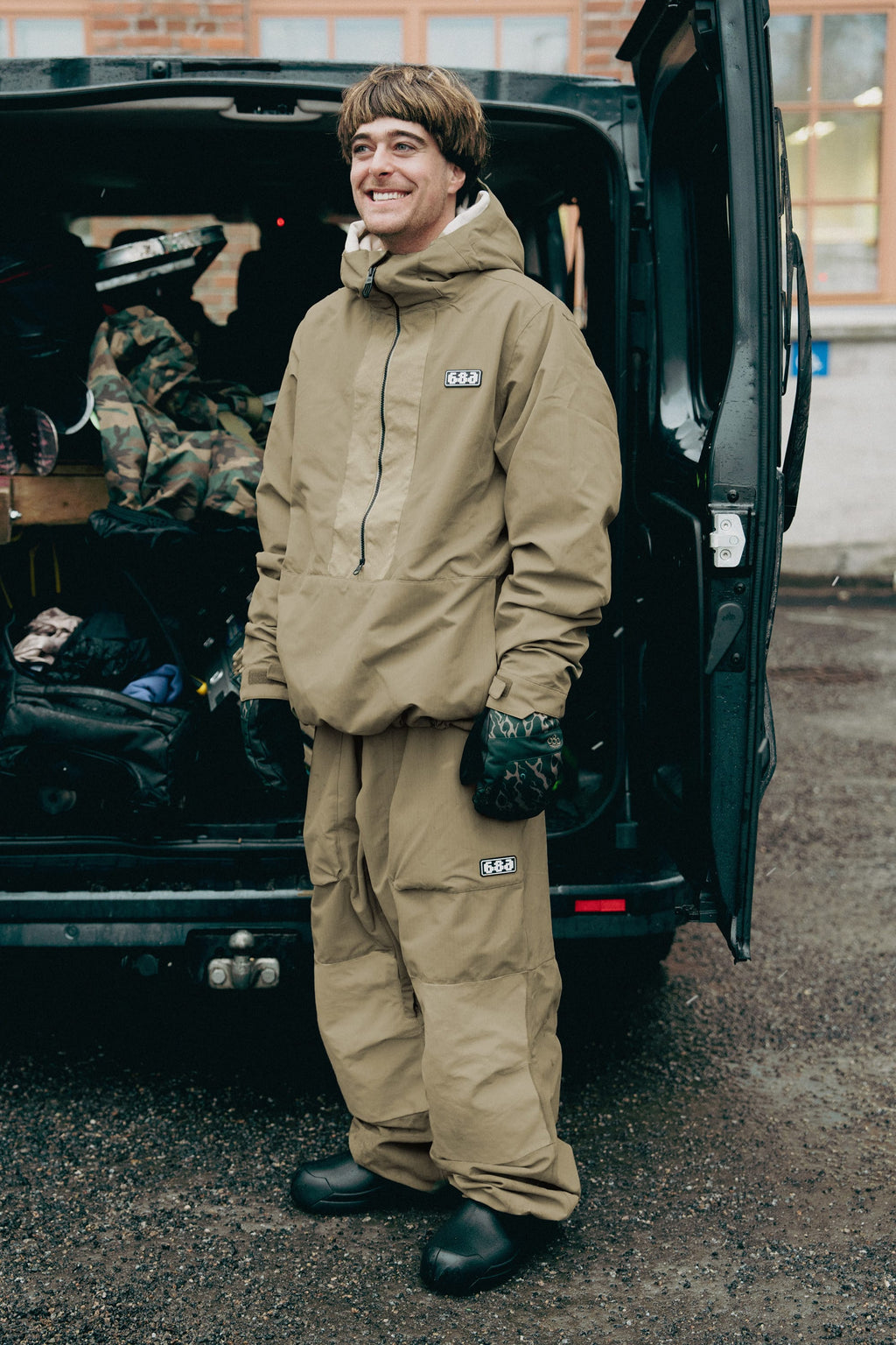 Wearing the 686 Ghost™ 2.5L Pant, a person stands smiling by an open van packed with gear on a wet surface, dressed in a tan hooded jacket, patterned gloves, and black shoes.