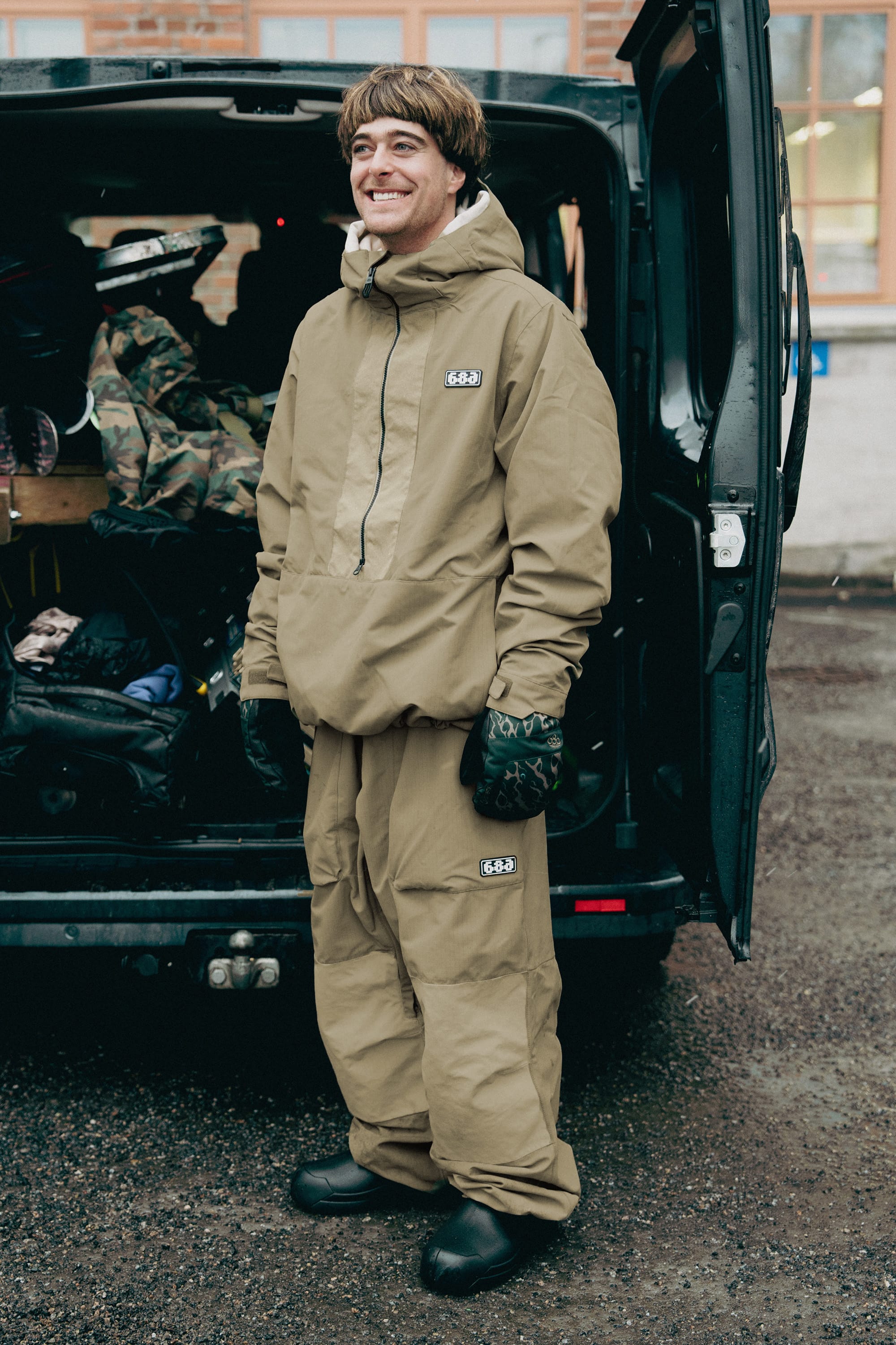 Wearing the 686 Ghost™ 2.5L Pant, a person stands smiling by an open van packed with gear on a wet surface, dressed in a tan hooded jacket, patterned gloves, and black shoes.