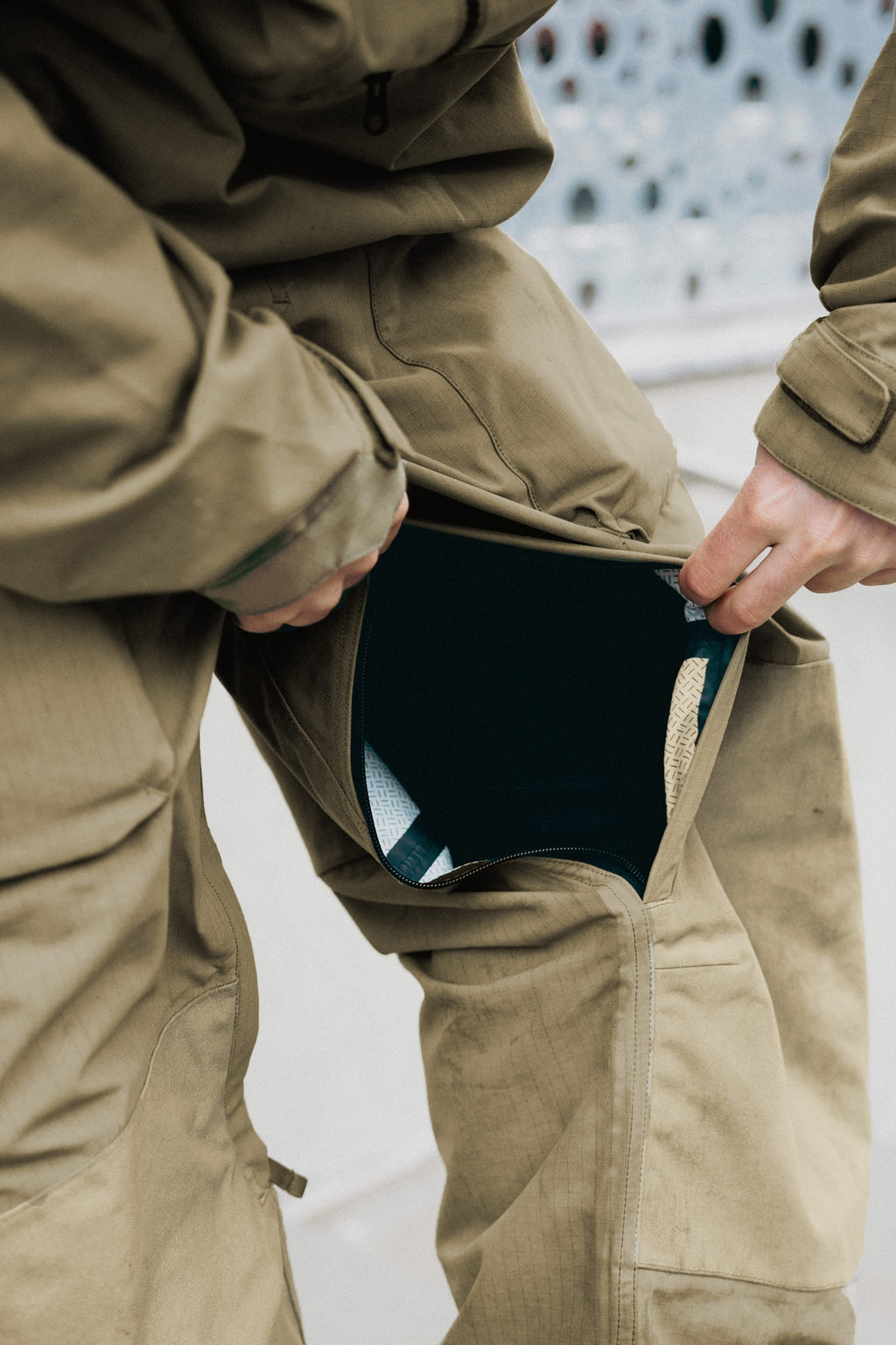 A person wearing the 686 Ghost™ 2.5L Pant opens a zippered side pocket, revealing a black inner lining with clothing underneath. Only their hands and part of the tan pants from 686 are visible.