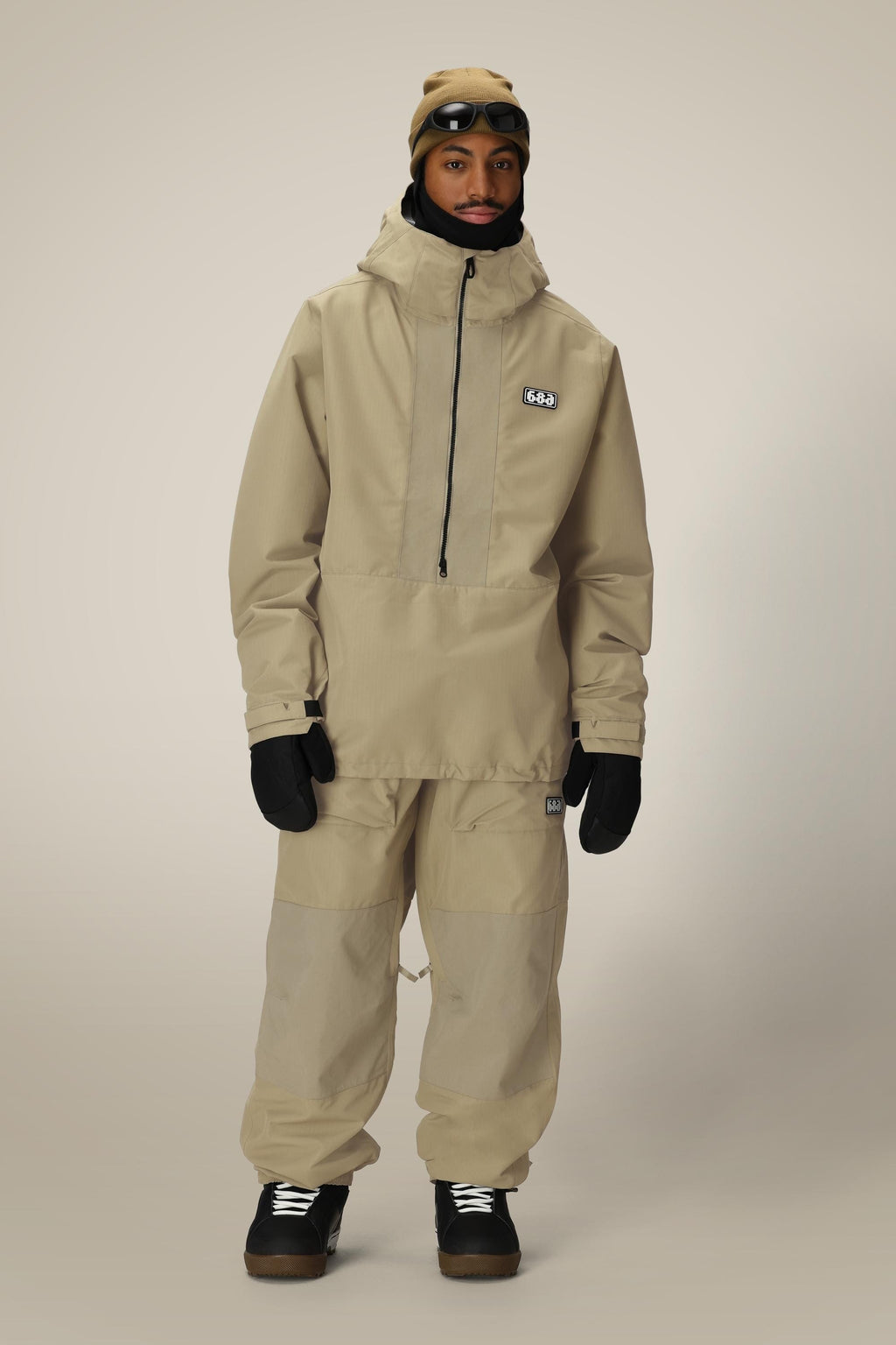A person wearing a beige hooded jacket, 686 Ghost™ 2.5L Pant by 686, black gloves and shoes, plus a beige beanie with goggles on top, stands against a plain light background.
