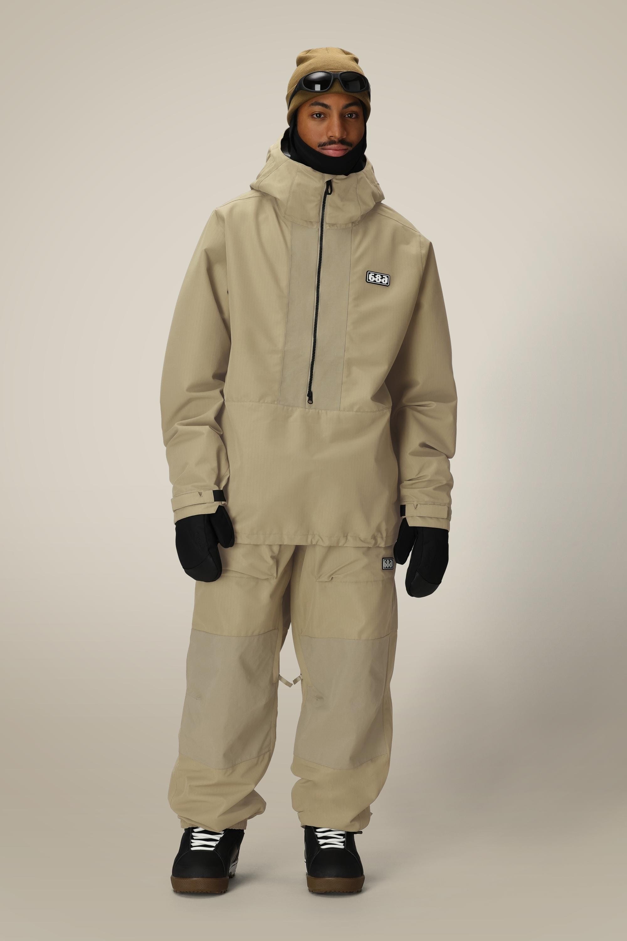 A person wearing a beige hooded jacket, 686 Ghost™ 2.5L Pant by 686, black gloves and shoes, plus a beige beanie with goggles on top, stands against a plain light background.