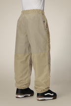 A person stands against a plain light background, seen from behind in relaxed-fit 686 Ghost™ 2.5L Pant by 686—loose-fitting, beige two-tone shell pants with elastic cuffs and waistband—paired with black and white sneakers.