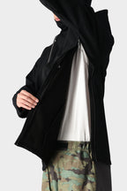 A person in camouflage pants and a white shirt unzips the black 686 Mens Waterproof Hoody, partially lifting their arm to reveal its fleece lining against a plain background.
