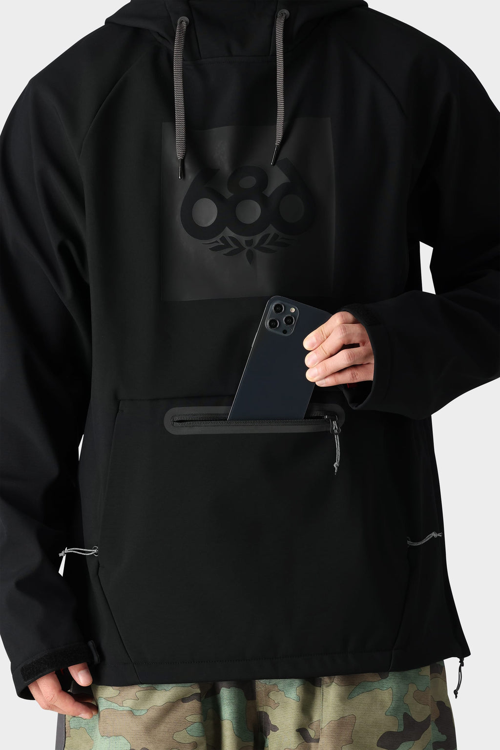 A person in the 686 Mens Waterproof Hoody, fleece-lined for comfort, with the 686 logo and front zippered pocket, pairs it with camo pants while placing a smartphone into the hoody’s pocket.