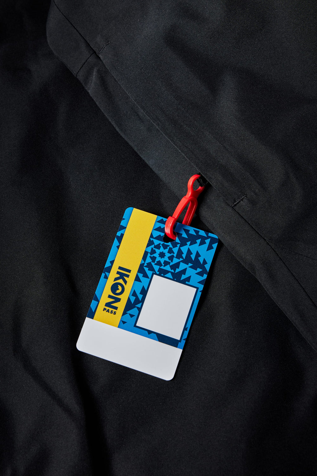 A ski pass with a blue and yellow design attached to a red lanyard clip rests on a black, fleece-lined 686 Mens Waterproof Hoody by 686. The pass displays IKON PASS and a pattern of blue triangles.
