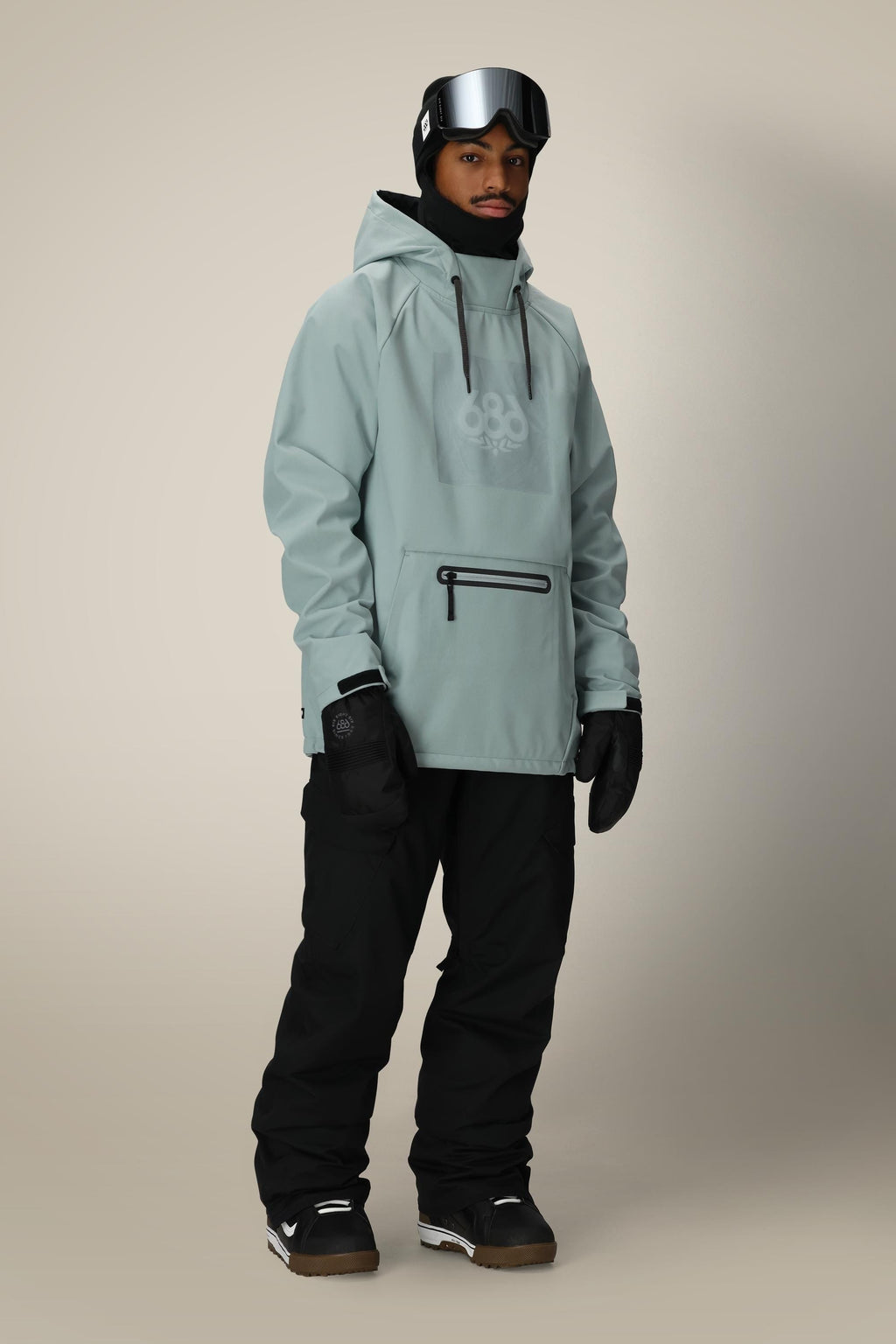 A person wears the 686 Mens Waterproof Hoody in light gray, paired with black snow pants, gloves, boots, a black beanie, and reflective ski goggles on their forehead—fully outfitted for winter sports.