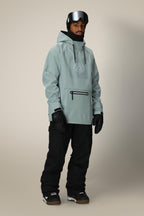 A person wears the 686 Mens Waterproof Hoody in light gray, paired with black snow pants, gloves, boots, a black beanie, and reflective ski goggles on their forehead—fully outfitted for winter sports.