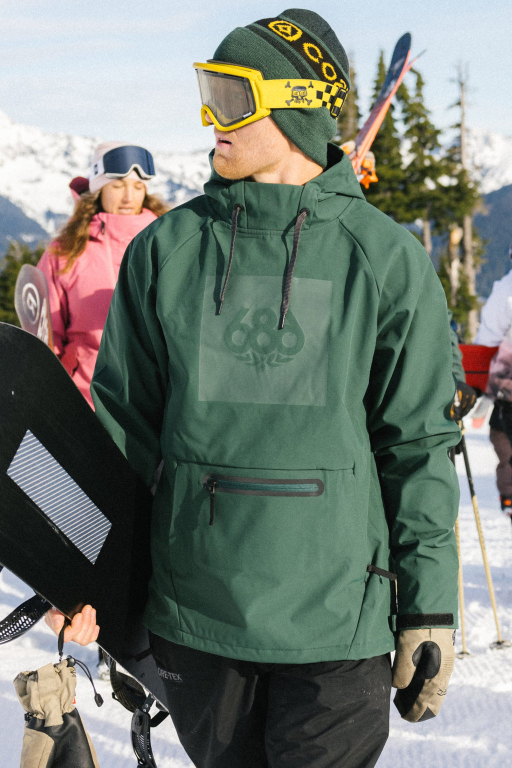 A man wearing the 686 Mens Waterproof Hoody stands on a snowy mountain holding a snowboard, while a woman in pink winter gear and snowboards appears in the background. Snowy peaks and pine trees are visible in the distance.