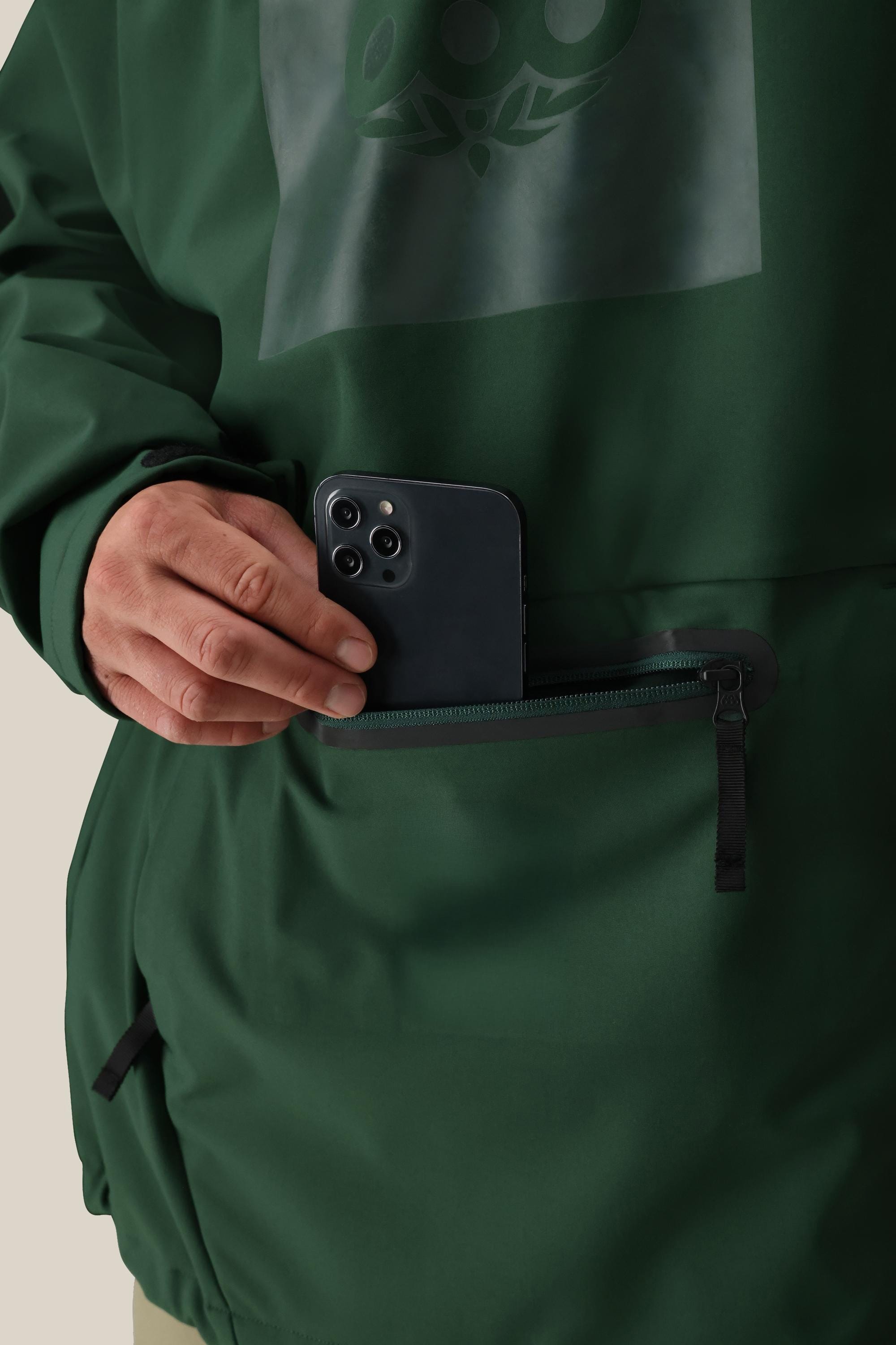 A person wearing the 686 Mens Waterproof Hoody in dark green places a black smartphone into its zippered front pocket.