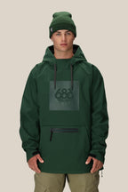 A man faces forward against a beige background wearing the 686 Mens Waterproof Hoody in dark green, featuring a black zipper, front pocket, and fleece-lined interior, along with matching khaki pants and a green 686 beanie.