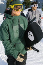 A snowboarder in a 686 Mens Waterproof Hoody and yellow goggles holds a black snowboard with a white skull graphic. Another person in winter gear stands on a snowy slope with trees in the background.