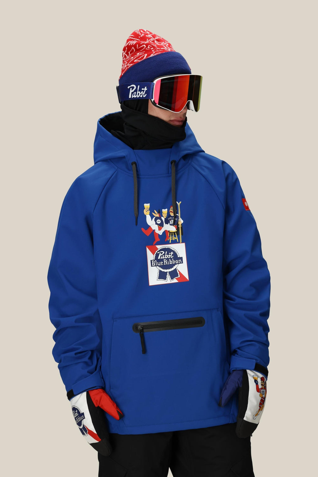 A person in a blue 686 Mens Waterproof Hoody, red and white beanie, ski goggles, and winter gloves stands against a neutral background, looking to the side. The hoody features a festive graphic and the 686 brand logo.