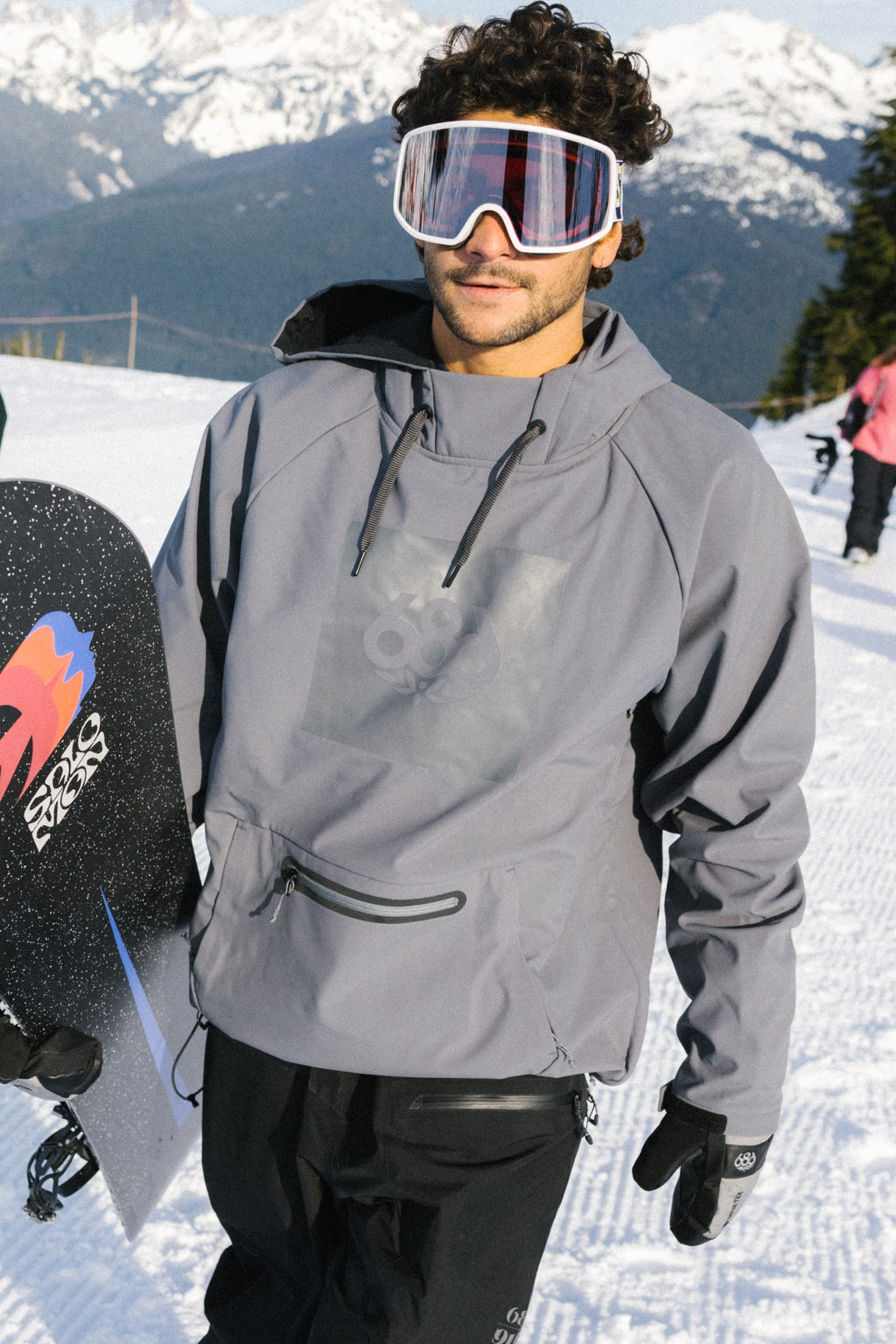 A man in a 686 Mens Waterproof Hoody and ski goggles stands on a snowy mountain slope with a snowboard. Snow-covered mountains and another person in winter gear appear in the background.