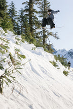 A snowboarder wearing the 686 Mens Waterproof Hoody by 686 catches air off a snowy slope, surrounded by evergreens and mountains under a bright, clear sky.