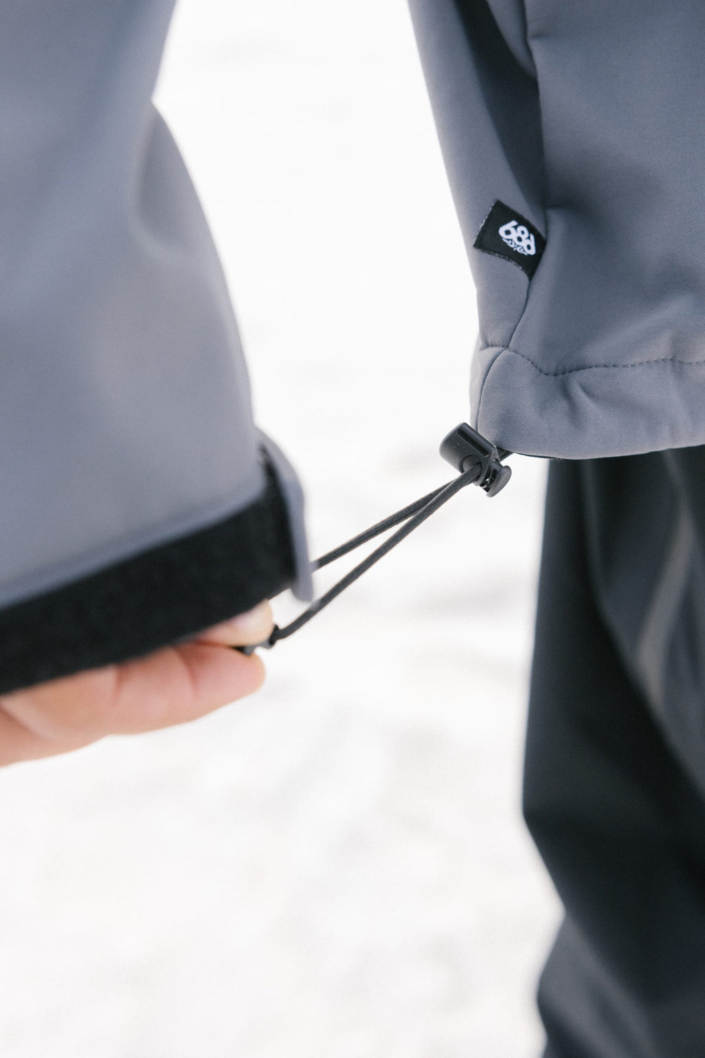 A close-up shows someone adjusting the elastic cuff drawstring on a gray 686 Mens Waterproof Hoody sleeve, fleece lined with a visible black and white 686 logo. The background is blurred and light-colored.