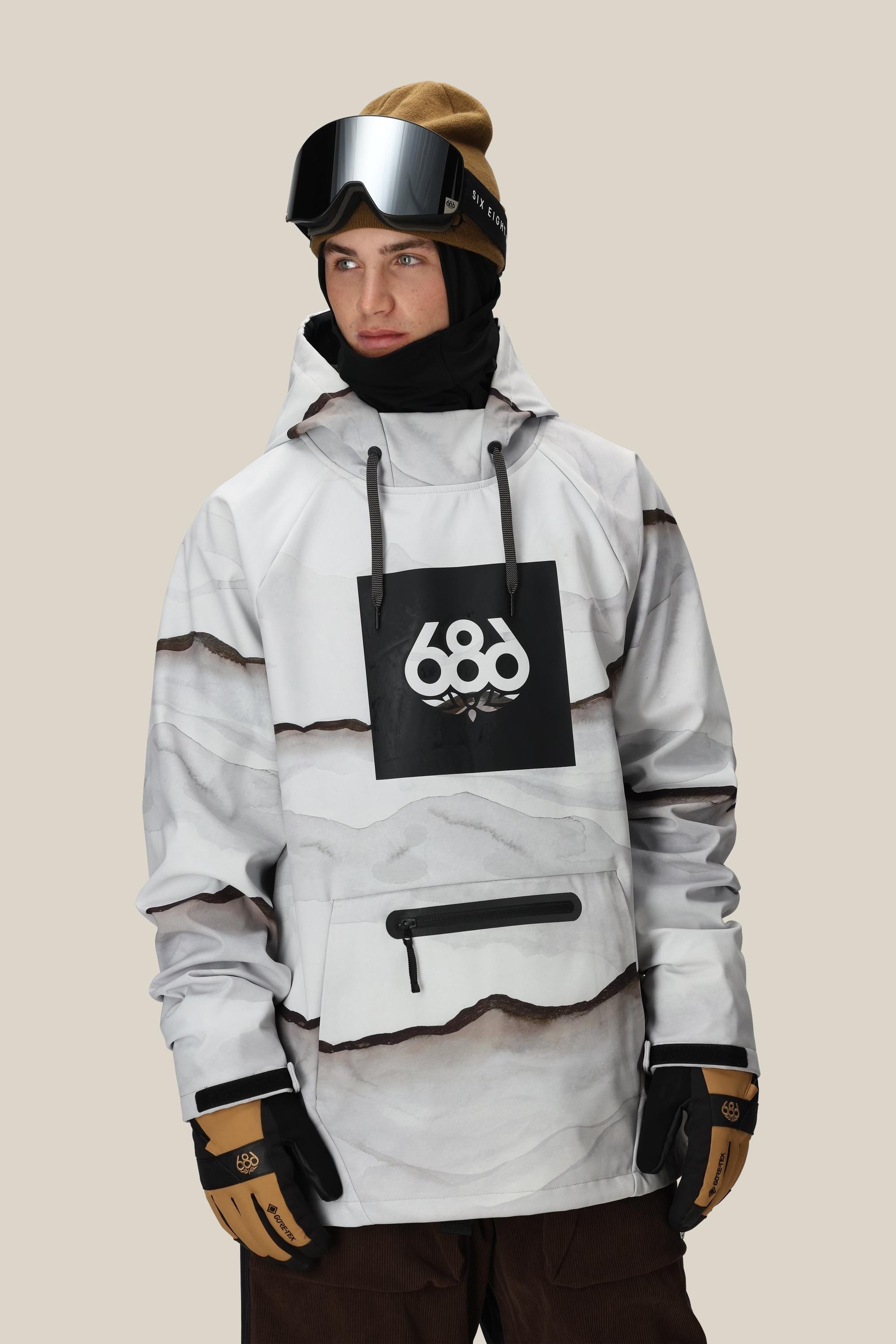 A person wearing the 686 Mens Waterproof Hoody with a black 686 logo, tan gloves, brown pants, a brown beanie, black ski goggles, and a black neck gaiter stands against a plain background.