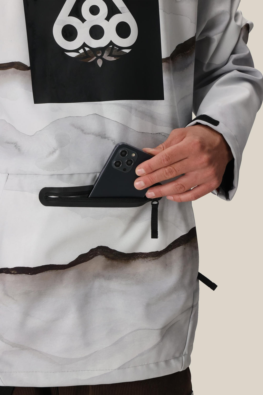 A person wearing a 686 Mens Waterproof Hoody in light gray and white pattern with a black 686 logo places a black smartphone into the front zippered pocket. Only their torso and hands are visible.