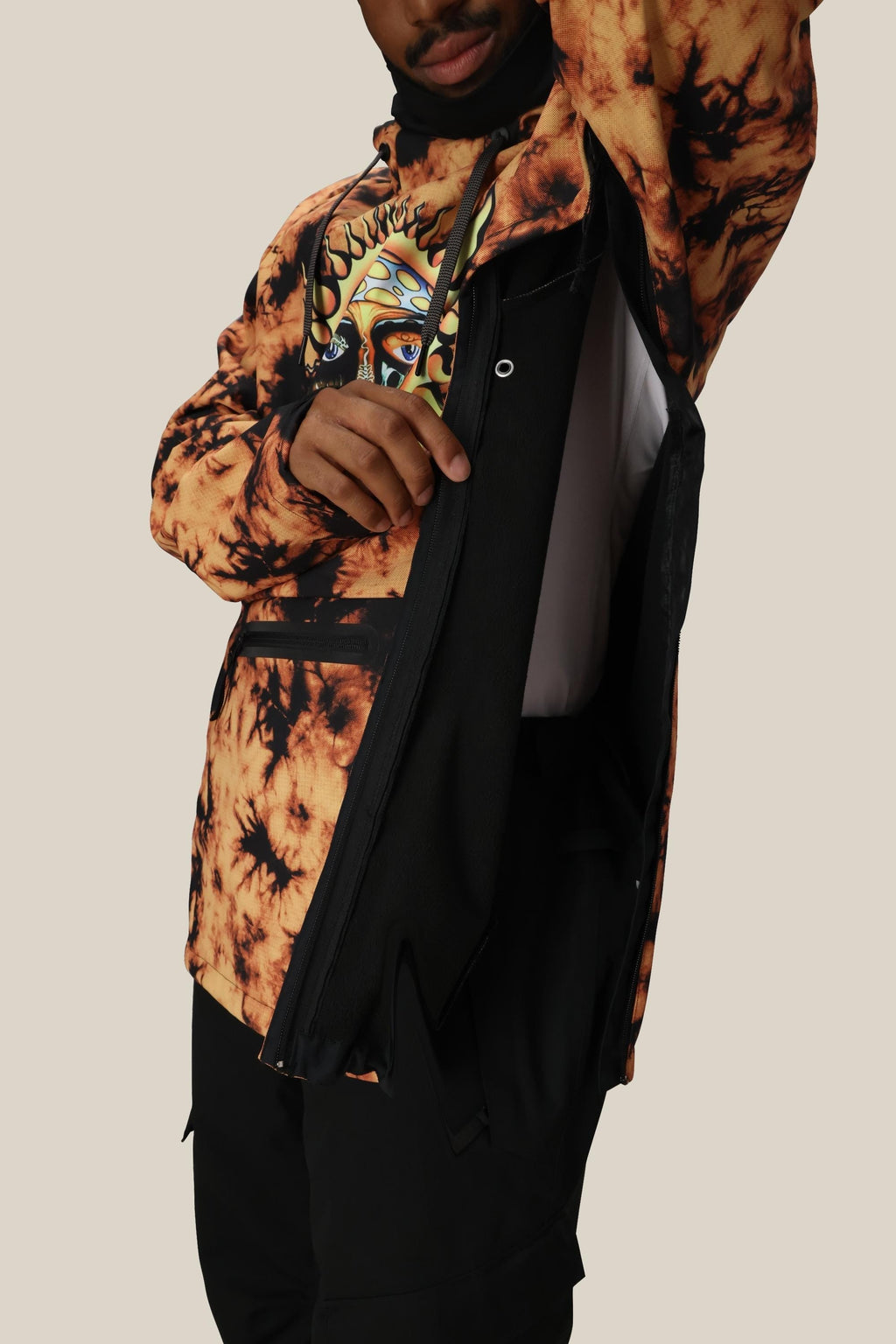 A person wearing the 686 Mens Waterproof Hoody in tie-dye orange and black with a graphic tee lifts one arm, revealing a side zipper and black fleece lining. The plain, light background highlights the 686 brand hoody’s features.