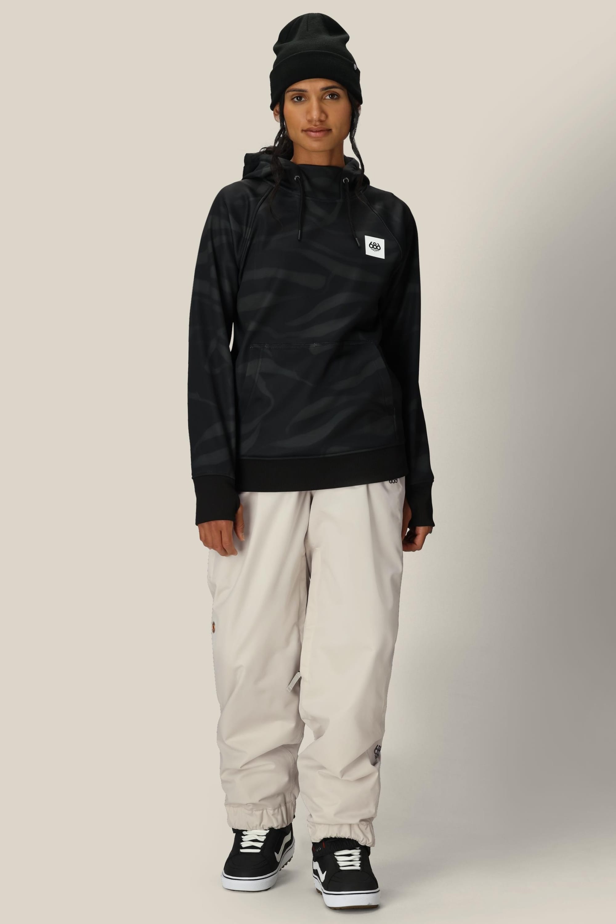 A person in a black beanie, beige baggy pants, black sneakers, and the 686 Womens Bonded Fleece Pullover Hoody by 686 stands facing forward against a light background.