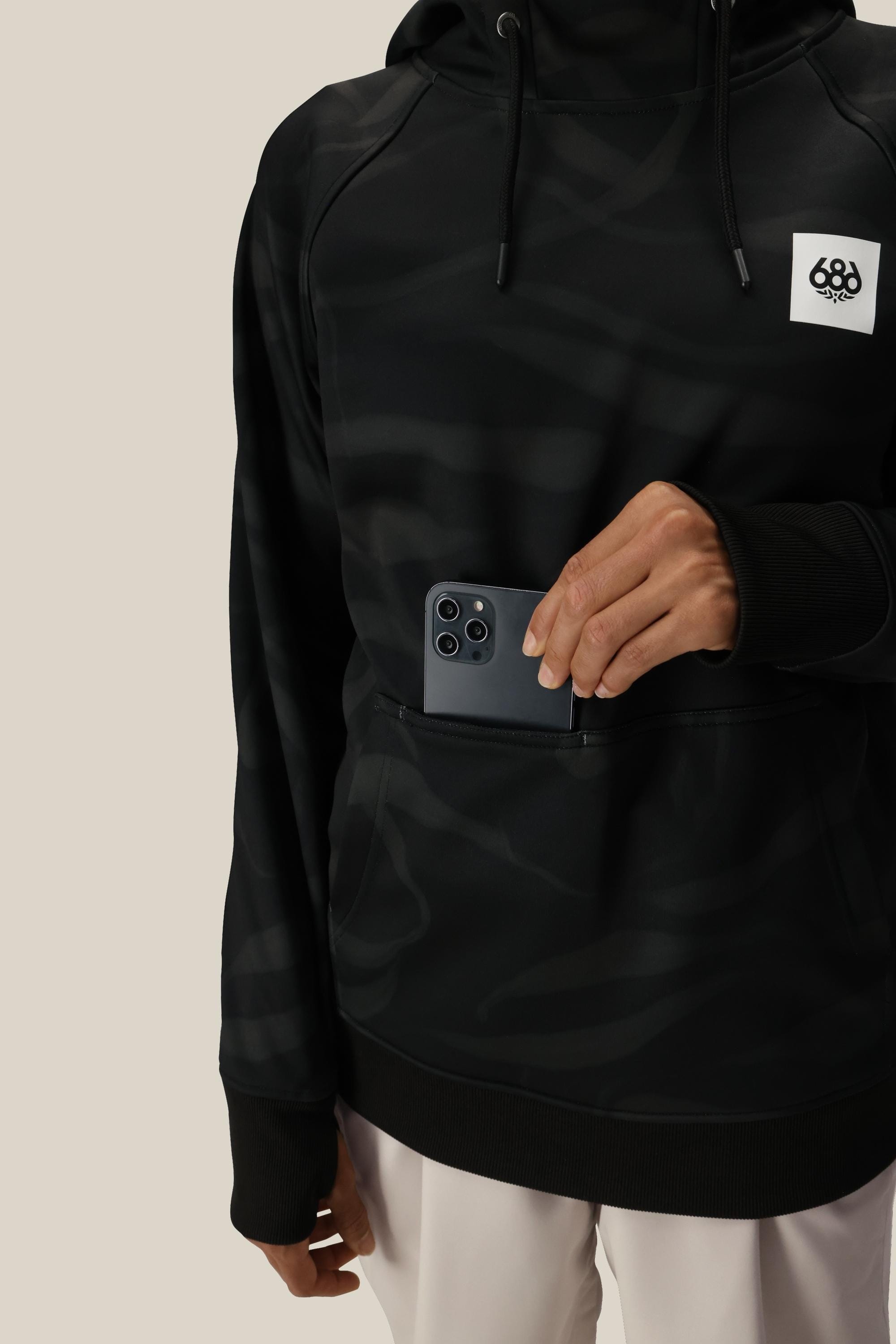 A person wearing the 686 Womens Bonded Fleece Pullover Hoody in black, showing only their torso and hand, places a black smartphone into the front pocket. The hoody features a small white logo; the background is plain beige.