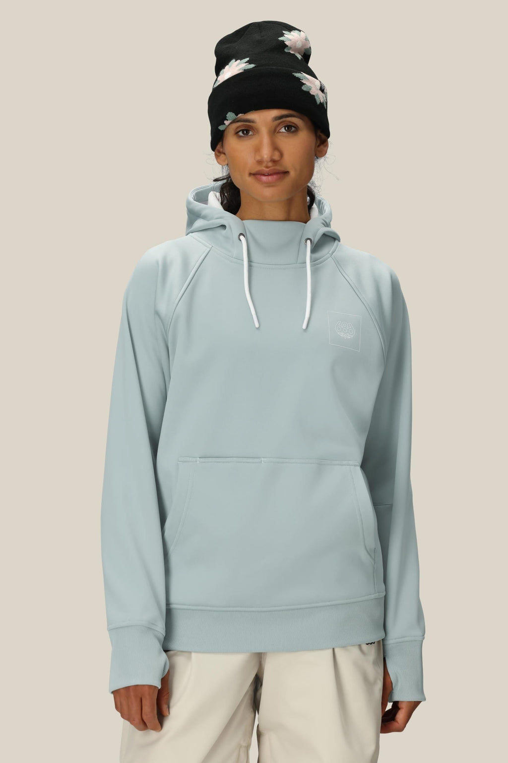 A person wears the 686 Womens Bonded Fleece Pullover Hoody in light blue with white drawstrings, paired with off-white pants and a black floral beanie, standing against a plain beige background.