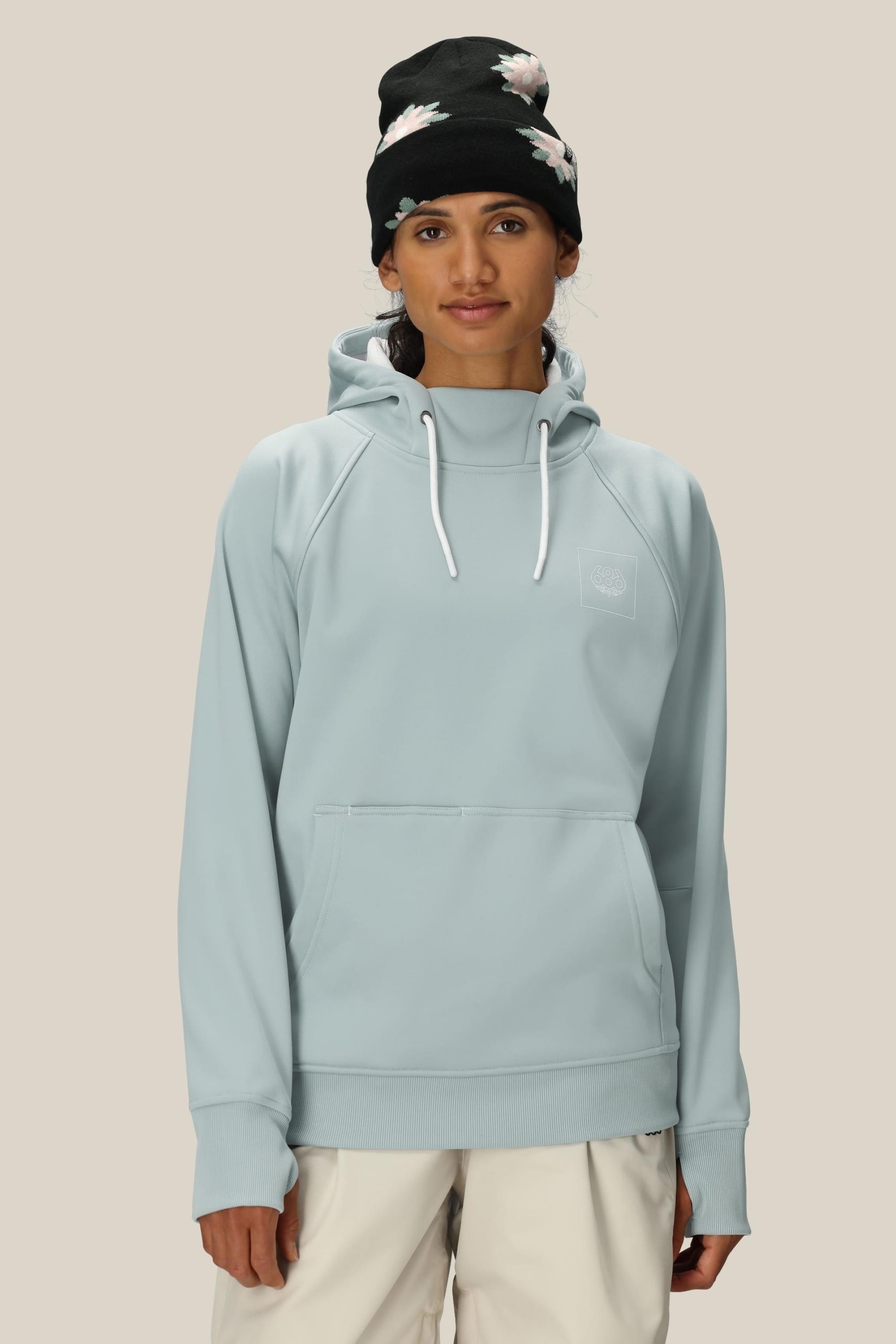 A person wears the 686 Womens Bonded Fleece Pullover Hoody in light blue with white drawstrings, paired with off-white pants and a black floral beanie, standing against a plain beige background.
