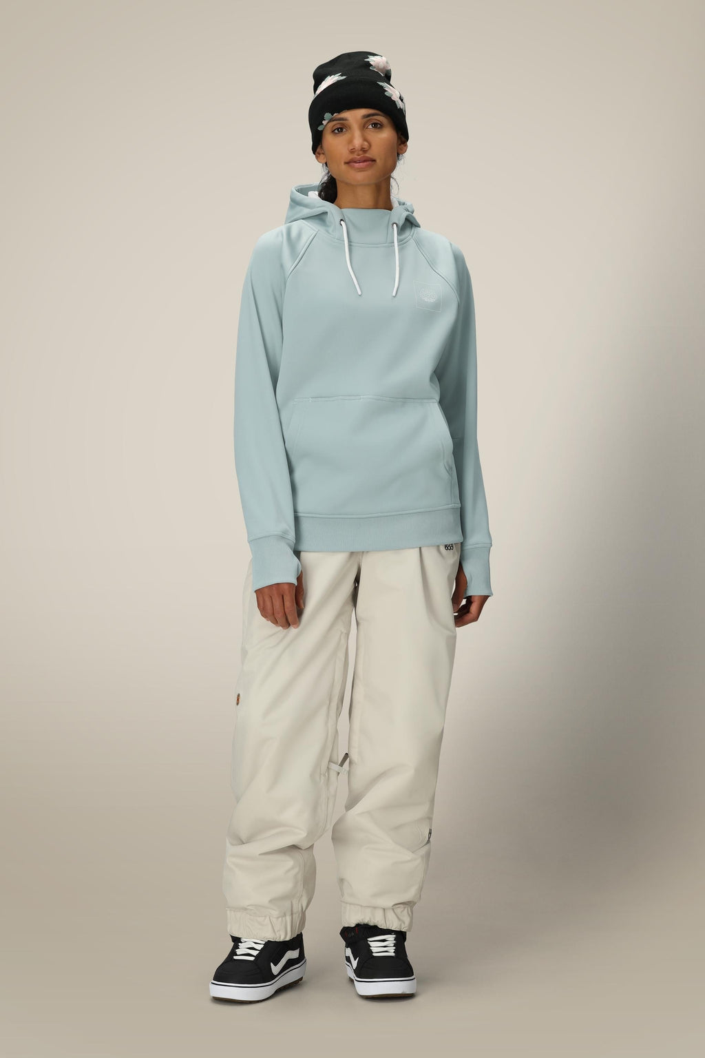 A person stands facing forward against a plain, light background, wearing the 686 Womens Bonded Fleece Pullover Hoody in light blue, off-white baggy pants, black Vans sneakers, and a black beanie with white accents.