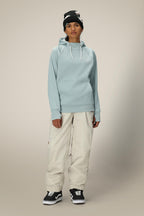 A person stands facing forward against a plain, light background, wearing the 686 Womens Bonded Fleece Pullover Hoody in light blue, off-white baggy pants, black Vans sneakers, and a black beanie with white accents.